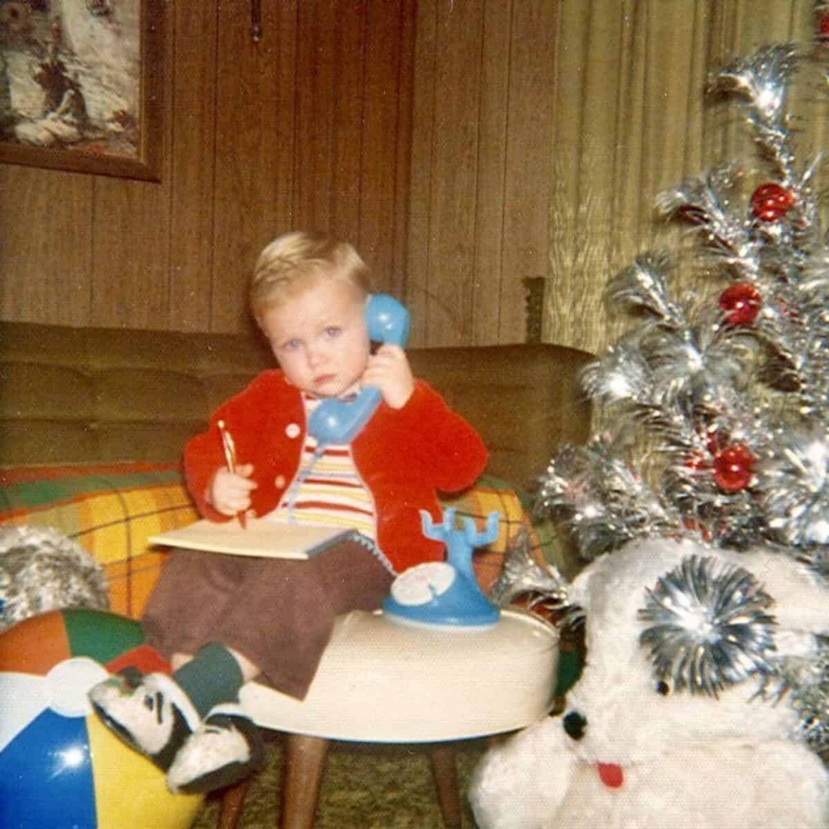 Child talking on a vintage blue rotary phone with a notepad, sitting on a colorful couch next to a silver Christmas tree with red ornaments, in a cozy retro living room.